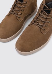 Botas de tobillo de ante marrón con una textura suave, forma de punta redonda, suela de goma color caramelo y cordones marrones. Presentan ojales metálicos para el cordón.