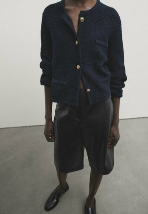 Personne portant un cardigan boutonné bleu marine texturé avec des boutons dorés, un short en cuir noir jusqu'aux genoux et des chaussures noires faciles à enfiler.