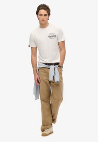 Witte crewneck t-shirt met zwarte graphic, beige corduroy broek en een blauw gestreepte shirt om de taille geknoopt. Tan sneakers maken de look compleet.