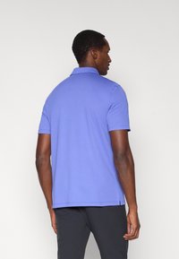 Vijolična polo majica s standardnim ovratnikom, kratkimi rokavi in stranskimi razporki na spodnjem robu. Gladek tekstura tkanine. Brez vidnih logotipov ali vzorcev.