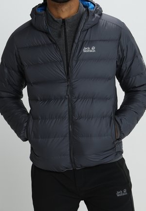 Homme portant une veste matelassée gris foncé de Jack Wolfskin avec les mains dans les poches et un pantalon noir assorti de Jack Wolfskin.