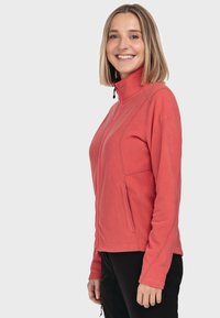 Fleece-Jacke in korallenpink mit hohem Kragen, Reißverschluss, seitlichen Taschen und schlankem Design. Sanfte Textur und figurnahe Silhouette.