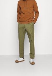 Felpa marrone con tasca frontale, pantaloni verdi con caviglie risvoltate e sneakers bianche con accenti marroni, caratterizzati da un design semplice.