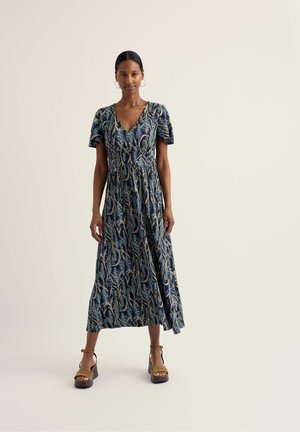 Žena stojaca v tmavomodrých midi šatách s listovým vzorom, krátkymi rukávmi, výstrihom do V a hnedými platformovými sandálmi na jednoduchom pozadí.