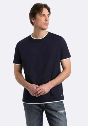 Uomo con camicia blu scuro a maniche corte con bordo bianco, in piedi con le mani intrecciate, guarda verso destra su uno sfondo chiaro uniforme.