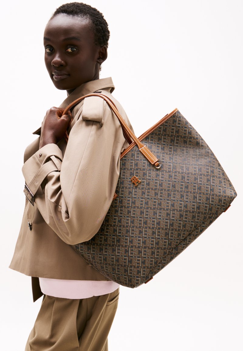 Geometrikus mintázatú szövet táska szürke és barna színekben, világosbarna bőr fogantyúkkal és díszítésekkel. A táska tágas, téglalap alakú.