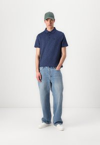 Camisola polo azul-marinho com uma textura subtil, combinada com jeans baggy azul-claro. Os acessórios incluem um boné verde e ténis brancos.