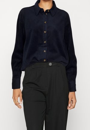 Mujer que lleva una camisa de pana azul marino oscuro abotonada, metida dentro de pantalones negros de talle alto con cierre de un solo botón.