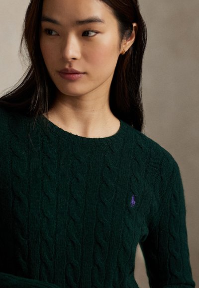 Giovane donna con capelli lunghi e scuri che indossa un maglione a trecce verde scuro con un piccolo logo ricamato viola, guarda verso sinistra.