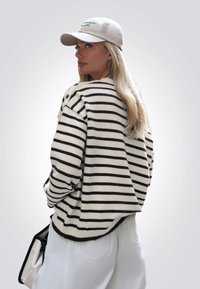 Maglione a righe in bianco e nero, taglio oversize, con scollatura rotonda e dettagli con bottoni. Indossato con un berretto beige e pantaloni bianchi per creare contrasto.