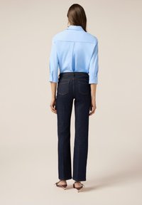 Camisa de botón azul claro con mangas tres cuartos, combinada con jeans oscuros de cintura alta. Tejido suave, corte clásico y líneas limpias.