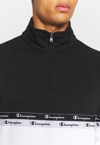 Felpa nera con zip e colletto alto. Presenta una banda di design bianca con il logo "Champion" sul petto. Tessuto morbido.