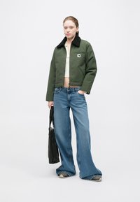 Groene cropped jas met zwarte fleece kraag, gecombineerd met lichtblauwe wijde jeans en een zwarte draagtas met zichtbare stikdetails.