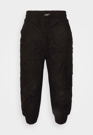 Hihaton musta paita edessä laskoksilla, mustat cargo jogger-housut, mustat sukat ja punaiset kuviolliset nahkakengät seisovalla henkilöllä.