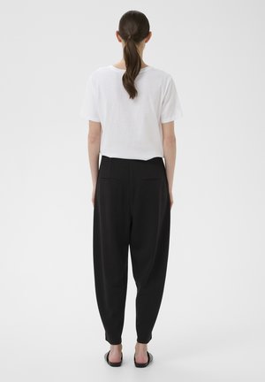 Femme aux cheveux bruns attachés en queue de cheval, portant une chemise blanche à manches courtes, un pantalon noir ample et des sandales noires à enfiler, debout devant un fond uni.