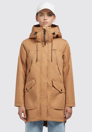 Braune wasserdichte Jacke mit Kapuze, Zugbandverschluss, Fronttaschen und Reißverschlussträger, kombiniert mit hellblauen Jeans.