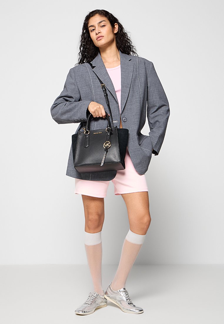 Borsa a tracolla nera in pelle con dettagli in oro, forma strutturata e un portachiavi. Il modello indossa un blazer grigio oversize e pantaloni corti rosa. Sneakers argentate.