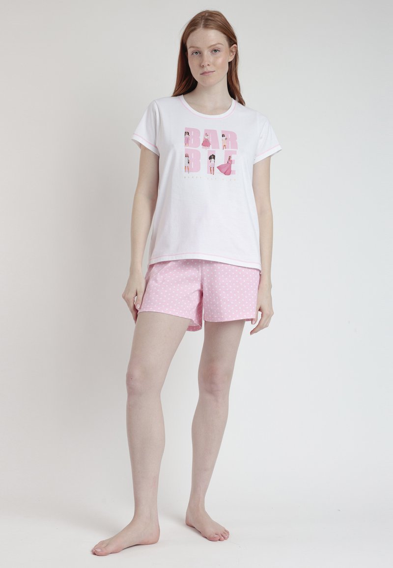 Joven mujer con cabello rojo vistiendo una camiseta blanca de manga corta con estampado de Barbie y pantalones cortos rosa con lunares, de pie descalza sobre un fondo liso.