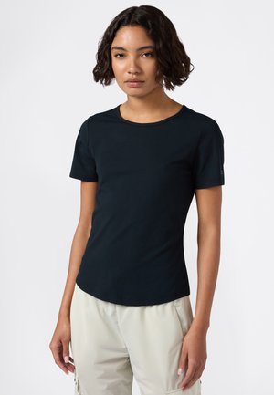 Donna con capelli ricci corti che indossa una maglietta nera a maniche corte e pantaloni cargo beige chiaro, in piedi su uno sfondo bianco.