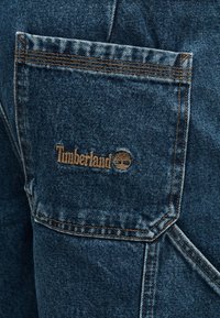 Μπλε τζιν τσέπη με καφέ λογότυπο Timberland και έμβλημα δέντρου κεντημένο στο κέντρο, λεπτομερειακά με πορτοκαλί ραφές.