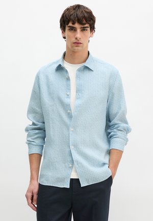 Młody mężczyzna ubrany w jasnoniebieską koszulę na guziki z wzorem, założoną na białą koszulkę, oraz ciemne spodnie, stojący na tle jednolitego białego tła.