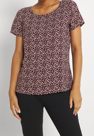 Vrouw die een bordeauxrode top met korte mouwen draagt, bedrukt met een klein wit en roze bloemenpatroon, gecombineerd met een zwarte broek, tegen een effen achtergrond.