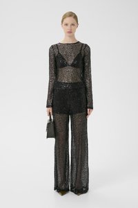Sort jumpsuit med pailletter, lange ærmer, gennemsigtigt stof og vide bukser, parret med en lille sort håndtaske.