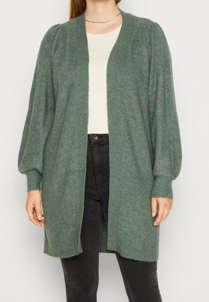 Kobieta w zielonym swetrze na drutach z przodu na otwarto, z bufkami na długich rękawach, białej bluzce, czarnych spodniach i cienkim złotym naszyjniku.