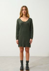 Abito di maglia verde con scollatura a V, maniche e orlo a costa. Abbinato a calze scure a coste e zoccoli neri su un pavimento neutro.