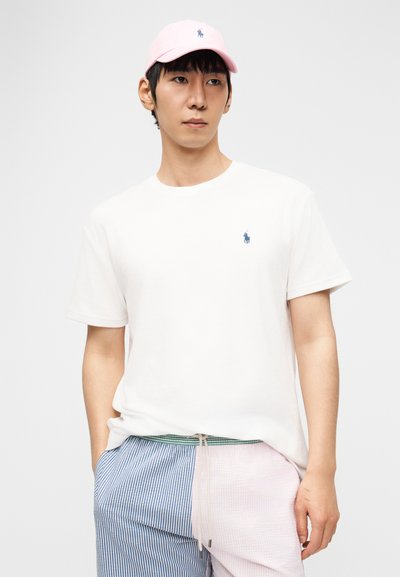 Νέος άνδρας που φοράει λευκό μπλουζάκι, ροζ καπέλο και δίχρωμο σορτς με κάθετες ρίγες σε μπλε και ροζ, στέκεται μπροστά σε απλό φόντο.