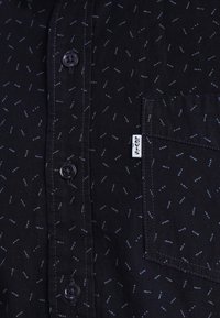 Svart skjorta med ett subtilt mönster av ljusblå prickar. Har knappar längs fram och en vänster bröstficka med ett märkesetikett.