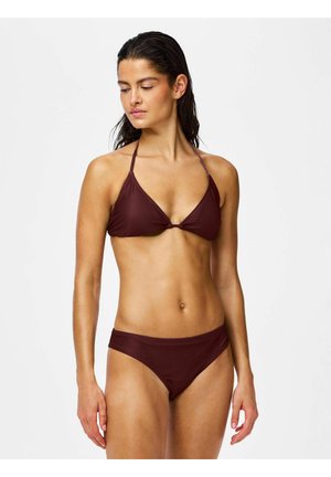 Frau mit nassen Haaren, die ein dunkelbraunes Neckholder-Bikinioberteil und passende Bikinihöschen vor einem einfarbigen hellen Hintergrund trägt.