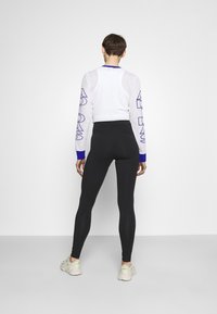 Bílý dlouhý rukáv top s modrými akcenty a detailem loga, spárovaný s černými legínami s vysokým pasem a světlými sportovními botami.