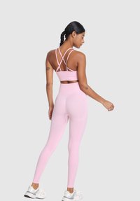 Ženska s kitkami, oblečena v svetlo roza športni modrček z navzkrižno vezanim hrbtom in hlače s visokim pasom, stoji v pozi v treh četrtinah obrnjena nazaj na sivi ozadju.