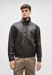 Pruunist nahast jakk kõrge kraega, ees nupuga sulgemine ja sile tekstuur. Jakk on voolav disainiga ja sisaldab peeneid õmbluse detaile.