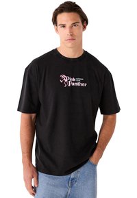 Črna bombažna majica s svetlo rožnato grafiko in napisom "Pink Panther Running Club." Dizajn je priložnostnega stila s kratkimi rokavi in sproščenim prileganjem.