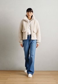 Beige faux karusnahk jope kõrge kraega ja kahe esipocketiga, kantud valge särgi ja siniste laiapõhjaliste teksade peal, kombineeritud valgete sportlike jalanõudega.