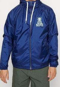 Marinblå, glänsande vindjacka i nylon, med en vit dragkedja och snoddar, samt en vit logotyp på bröstet.