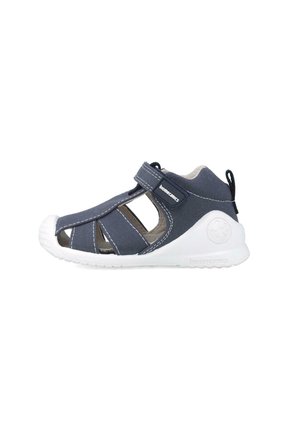 Chanclas de tela azul oscuro con suelas de goma blanca, que cuentan con lados abiertos, correa ajustable y puntera reforzada para mayor durabilidad.