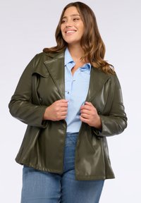 Olivgrön fusk läderjacka med sned krage, dubbelknapprad och avslappnad passform, bärs över en ljusblå skjorta.