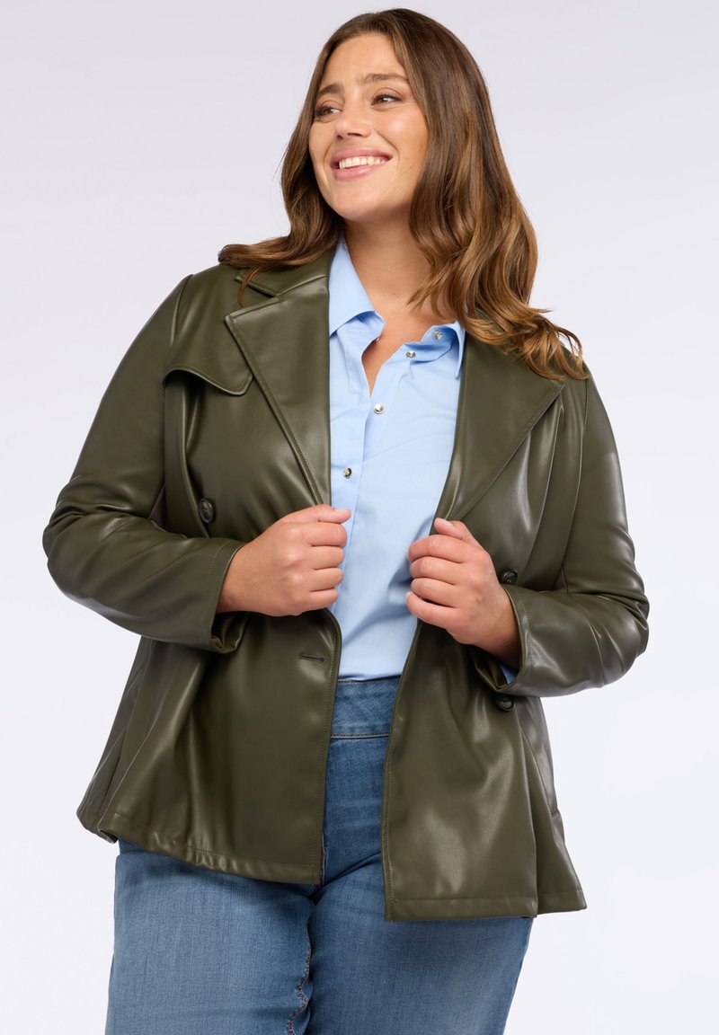 Olivgrön fusk läderjacka med sned krage, dubbelknapprad och avslappnad passform, bärs över en ljusblå skjorta.