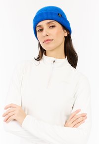 Blauwe gebreide beanie met een geribde textuur en een klein zwart label, gedragen over een witte rits-top. Focus op de levendige kleur en ontwerpdetails.