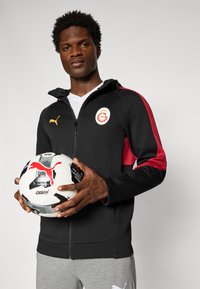 Musta vetoketjullinen takki maroon-vivahteilla ja kultaisella Puma-logolla. Harmaat fleece-housut, pitäen mustavalkoista jalkapalloa, jossa on Puma-brändi.
