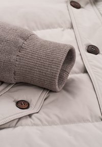 Daunenjacke mit einer glatten, hellgrauen Außenseite, einem strukturierten, beigen Strickbund und braunen runden Knöpfen.