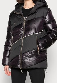 Giubbotto puffer nero con cappuccio, con pannelli diagonali lucidi e una zip argentata. Tessuto strutturato con sezioni lisce e opache.