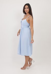 Vestido sin mangas de color azul claro, con un cuerpo ajustado, detalles de botones y una falda acampanada. Combinado con sandalias de tacón transparentes.