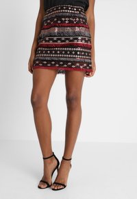 Benen in een zwart-rood geprint franje minirok, gecombineerd met zwart enkelband hoge hakken op een witte achtergrond.