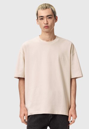 Jonge man met kort geblondeerd haar, die een loszittend lichtbeige T-shirt en donkere broek draagt tegen een effen lichte achtergrond.