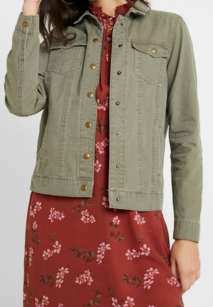 Vrouw draagt een olijfgroene jas met knopen over een rode bloemenjurk met roze en groene bloemenpatronen.