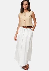 Beige bauchfreier Weste mit runden Knöpfen, kombiniert mit einem weißen Maxirock mit Falten und einem braunen Gürtel mit einer silbernen Schnalle. Braune Sandalen.